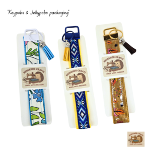 A collection of five wristlet keychains labeled "KeyPobs" and "JellyPobs" in various designs: geometric blue, floral blue, pink rose, iridescent jelly, and gold jelly with feather prints. Each has a matching metal clasp and tassel charm.