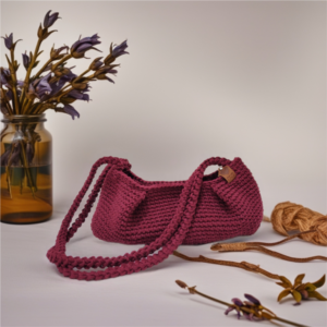 Handmade burgundy dumpling-style crochet bag with double braided straps, set with dried flowers and yarn on a neutral background.