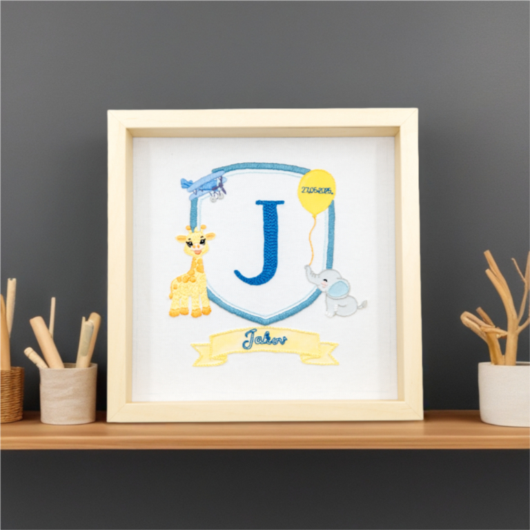 Framed embroidered artwork featuring a large blue letter "J" inside a blue shield. A yellow giraffe stands on the left, a small blue airplane flies above, and a gray elephant holds a yellow balloon on the right with the date stitched on it. Below the shield, a yellow banner displays the name in blue script. The piece is set in a light wooden frame on a white fabric background.