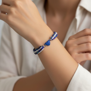 A handmade beaded bracelet with blue, white, and purple seed beads arranged in three strands, featuring a small blue heart-shaped clasp, worn on a woman's wrist.