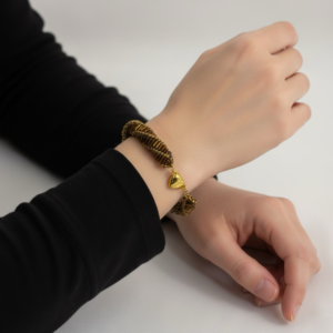 A close-up of a person wearing a handmade golden-brown woven bracelet with a small gold heart charm.