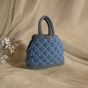 Handmade crochet handbag in grey with a textured blue geometric pattern, featuring sturdy double handles. Displayed on a neutral beige background with dried floral accents.