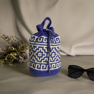 A handmade crochet bag with intricate blue and white geometric patterns, featuring a drawstring closure. The bag is placed next to a pair of black sunglasses and dried flowers, set against a soft neutral backdrop.