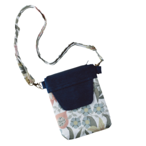 A small crossbody bag with a floral pattern in soft pastel colors. The bag has a flap made of dark blue fabric, possibly denim or canvas, and a matching top zipper. The strap is made of the same floral fabric as the bag and is attached with metal clasps.