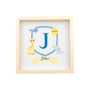 Framed embroidered artwork featuring a large blue letter "J" inside a blue shield. A yellow giraffe stands on the left, a small blue airplane flies above, and a gray elephant holds a yellow balloon on the right with the date stitched on it. Below the shield, a yellow banner displays the name in blue script. The piece is set in a light wooden frame on a white fabric background.