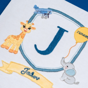 Framed embroidered artwork featuring a large blue letter "J" inside a blue shield. A yellow giraffe stands on the left, a small blue airplane flies above, and a gray elephant holds a yellow balloon on the right with the date stitched on it. Below the shield, a yellow banner displays the name in blue script. The piece is set in a light wooden frame on a white fabric background.